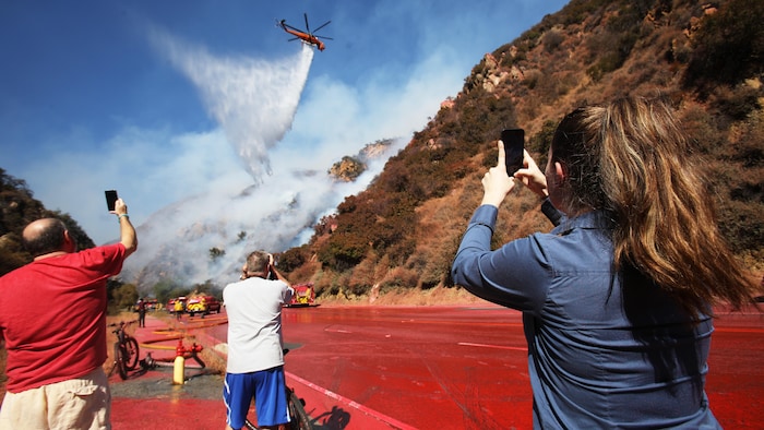 Des personnes prennent des photos d'un hélicoptère qui effectue un largage d'eau sur un feu de forêt menaçant une maison située à flanc de colline dans le quartier de Pacific Palisades, en Californie.