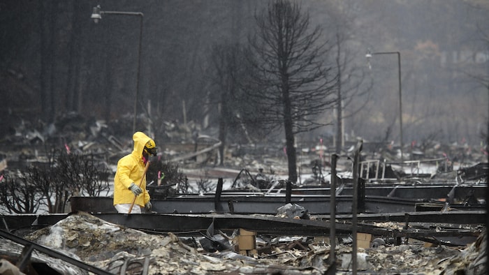 Une personne en veste jaune portant un masque à gaz marche dans les décombres d'un quartier détruit par un incendie.