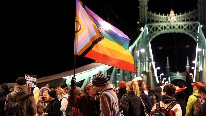 Deux personnes, une agitant un drapeau LGBT, s'embrassent durant une manifestation le soir, près d'un pont.