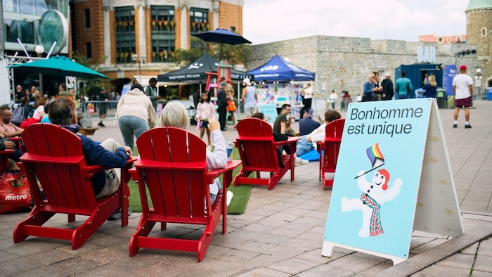 Public assis devant un panneau affichant Bonhomme Carnaval brandissant un drapeau de la Fierté.