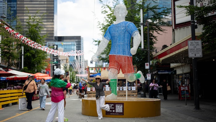 Des touristes prennent des photos devant une sculpture géante d'un joueur de soccer dans la rue piétonne Granville.