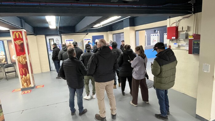 Une foule de personnes attend en file à l'intérieur d'un bâtiment aux murs beiges et plafonds bas. À gauche, un pilier publicitaire affiche des frites McDonald's. À droite, des boîtiers électriques rouges sont fixés au mur près d'une ouverture.