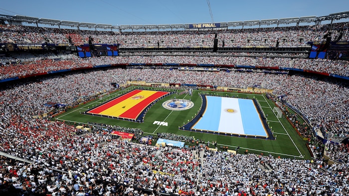 Des estrades sont bondées alors que deux immenses drapeaux de l'Espagne et de l'Argentine sont déployés sur le terrain de soccer, entourés des joueurs des équipes respectives.