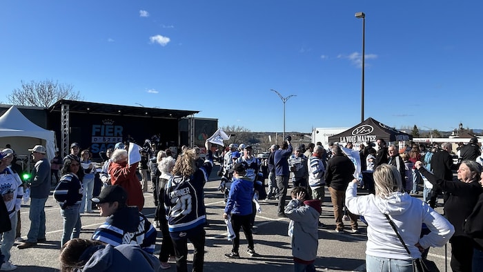 Une foule de personnes est rassemblée à l'extérieur dans un grand stationnement par une journée ensoleillée. À gauche, une scène affiche le slogan « Fier d'être Sags ». De nombreux spectateurs portent des chandails bleus et blancs aux couleurs de l'équipe.