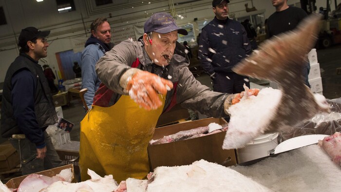 Un homme portant une casquette et un tablier jaune lance un gros poisson congelé sur une table, envoyant de la neige partout, au milieu d'un marché public. Quatre hommes derrière observent la scène.