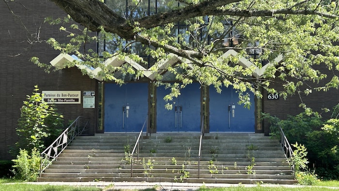 Le bâtiment de face. Des mauvaises herbes poussent dans les marches et la peinture des portes se détache.