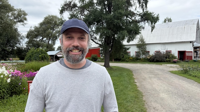 Un homme barbu portant une casquette bleue et un pull gris sourit à l'extérieur.