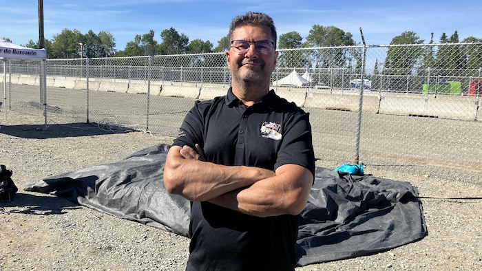 Éric Bourque, les bras croisés, pose pour la caméra devant une clôture en esquissant un léger sourire.