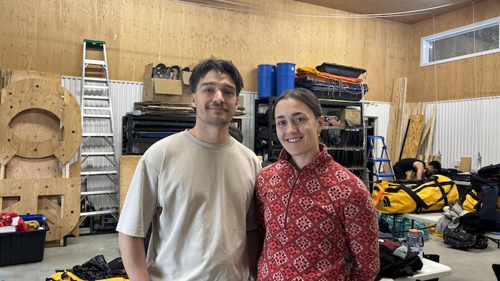 Un jeune homme et une jeune femme sourient face à la caméra dans un centre d'équipement de plein air. L'arrière-plan montre des étagères chargées, des échelles, des barils bleus et des sacs d'expédition jaunes sur des tables.