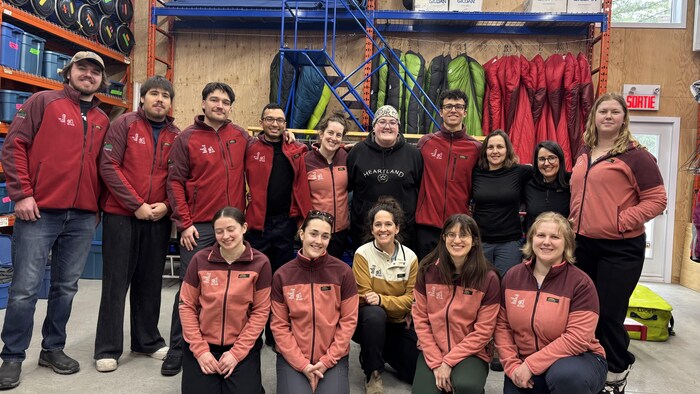 Un groupe de quatorze personnes pose pour une photo d'équipe dans un entrepôt de matériel. La majorité porte des vestes polaires rouges et bordeaux identiques. Ils sont entourés de sacs de couchage suspendus et de bacs de rangement.