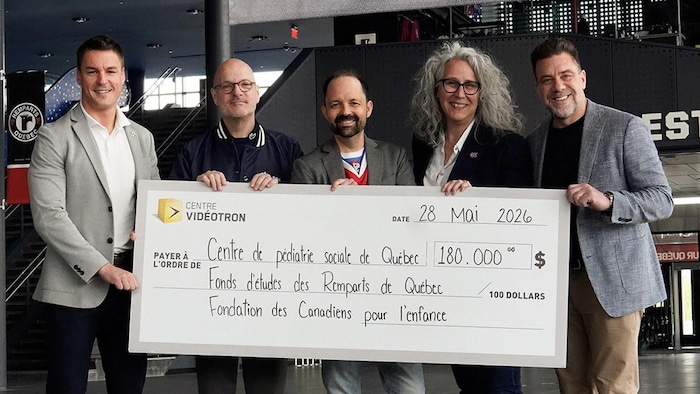 Tommy Castonguay, Claude Lavoie, Jean-Christophe Nicolas, Geneviève Paquette et Martin Tremblay tiennent un gros chèque daté du 28 mai 2026. Il est payé à l'ordre des Centre de pédiatrie sociale de Québec, du Fonds d'études des Remparts de Québec et de la Fondation des Canadiens pour l'enfance. Il sont dans l'atrium du Centre Vidéotron. 