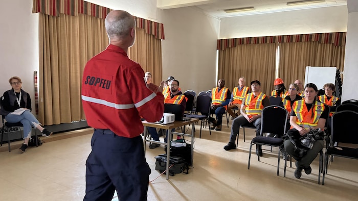 Un formateur portant une chemise sur laquelle le mot SOPFEU est écrit au dos. L'homme est debout, dos à la caméra, face à une dizaine de personnes assises, l'écoutant attentivement. Ils sont dans une salle où les rideaux sont fermés.