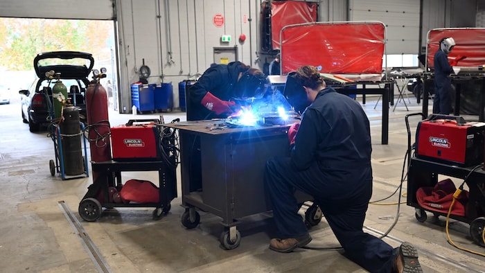 Deux personnes équipées de masques de protection effectuent des travaux de soudure sur des pièces métalliques dans un atelier de formation, tandis qu’une troisième travaille à l’arrière-plan.