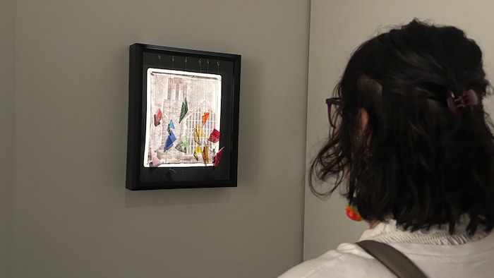 Plan de profil d'une personne avec des cheveux foncés et attachés, portant des lunettes, regardant une œuvre d'art encadrée suspendue sur un mur texturé de la galerie.