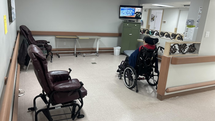 Une femme assise en fauteuil roulant écoute la télévision dans une petite salle.