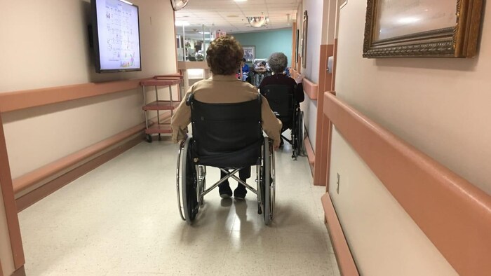 Deux dames âgées se déplacent en fauteuil roulant dans le couloir d'un foyer de soins.