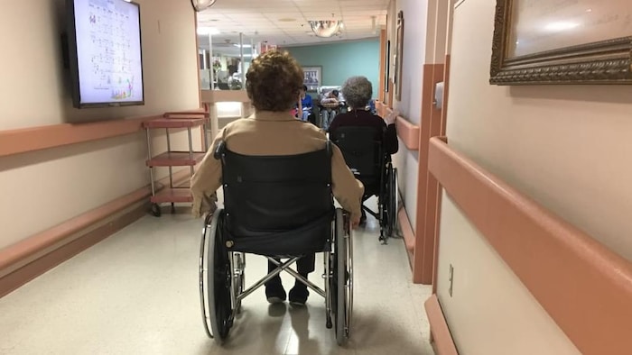 Deux dames âgées se déplacent en fauteuil roulant dans un couloir d'un foyer de soins.