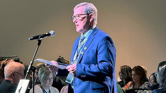 Un homme âgé aux cheveux gris, portant des lunettes et un complet bleu, se tient debout devant un microphone sur pied.