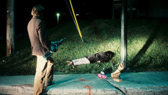 Un homme est debout avec un fusil de chasse emballé à côté d'un corps qui gît sur le bord du trottoir où est étalé un sac de provisions.