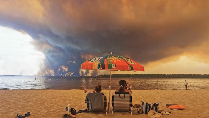Des baigneurs se reposent sur la plage de Moutchic, au bord de l'étang de Lacanau, à Lacanau, dans le sud-ouest de la France, le 24 juillet 2026, tandis que des nuages de fumée s'élèvent dans le ciel en raison de l'incendie qui ravage la Gironde.