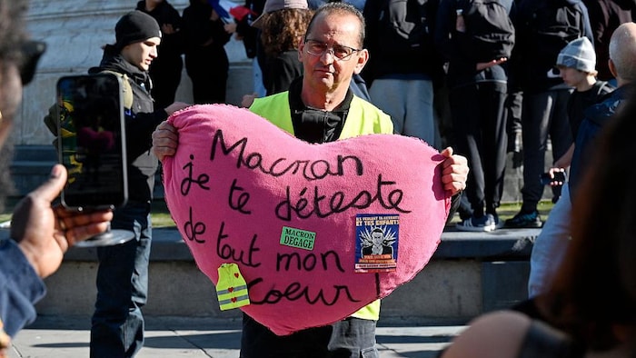 Un manifestant, vêtu d’un gilet jaune, tient un oreiller sur lequel on peut lire « Macron, je te déteste de tout mon cœur ».