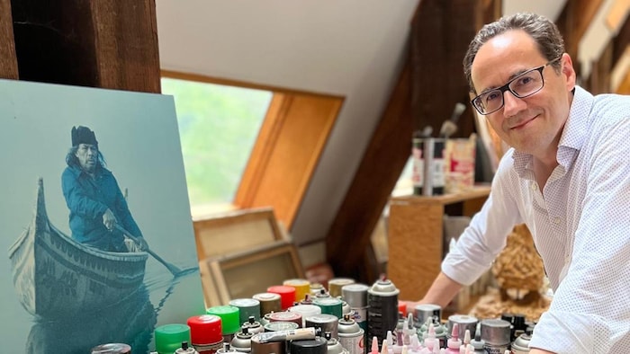 Un homme souriant dans un atelier pose la main sur une table garnie de nombreuses bombes aérosols et flacons de peinture.