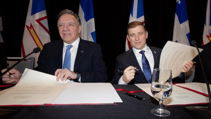 François Legault et Andrew Furey assis côte à côte avec des documents. Les deux tiennent des stylos à la main. Andrew Furey regarde la caméra en souriant, François Legault regarde ailleurs.