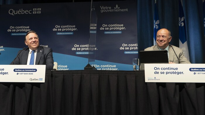 François Legault et Christian Dubé sont assis à la table de presse et sourient. 