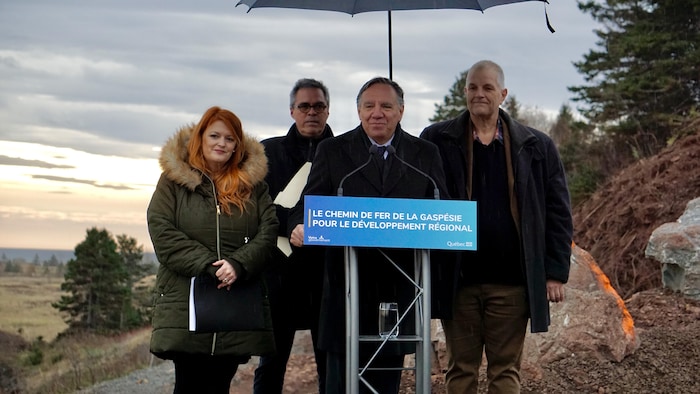 François Legault se trouve derrière un lutrin. Trois personnes se tiennent derrière lui.