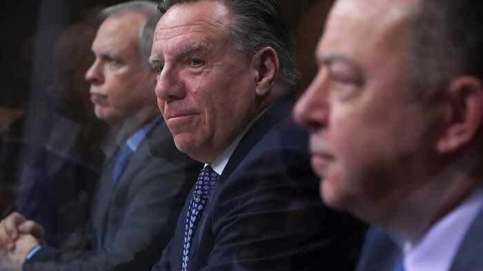 Luc Boileau (flou, à gauche), François Legault avec un léger sourire au centre et Christian Dubé (flou, à droite), à la table de la conférence de presse.