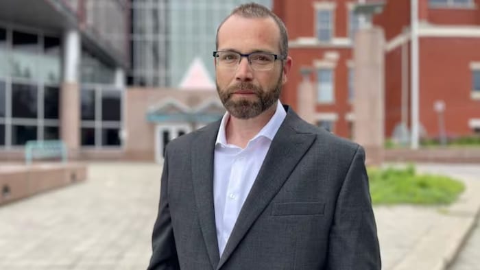 Un homme en veston fixe l'objectif, debout dans une place publique extérieure.