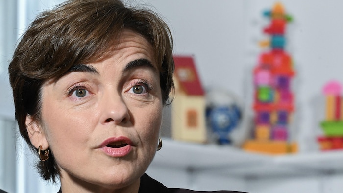 Christine Fréchette parle devant des jouets posés sur une étagère derrière elle.
