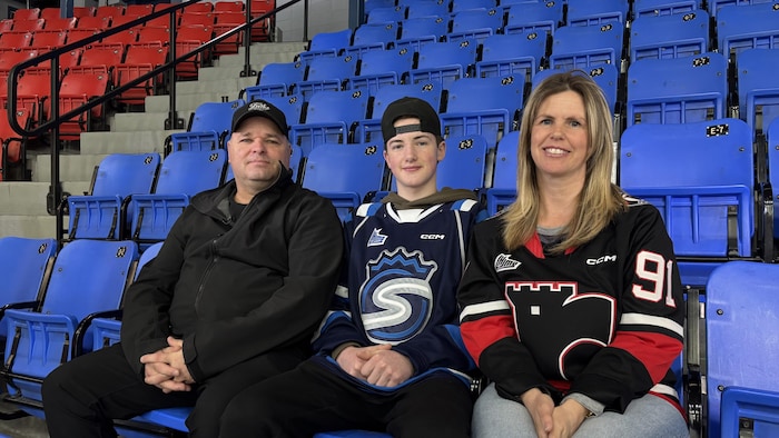 Trois personnes, un homme, un adolescent et une femme, posent assises dans les gradins bleus d’un aréna. Ils portent des vêtements de hockey, incluant des chandails de la LHJMQ des Saguenéens de Chicoutimi et des Remparts de Québec.