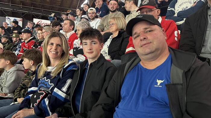 Une femme en chandail bleu des Saguenéens, un adolescent et un homme en t-shirt bleu et casquette noire posent assis dans les gradins. Ils sont entourés d'une foule de spectateurs assistant à un match de hockey dans un aréna.