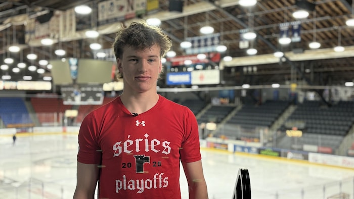 Un jeune homme aux cheveux bruns ébouriffés, vêtu d'un t-shirt rouge arborant l'inscription « séries 2026 playoffs » avec le logo des Remparts, se tient devant une patinoire de hockey. Les gradins sombres et le tableau indicateur sont visibles en arrière-plan.