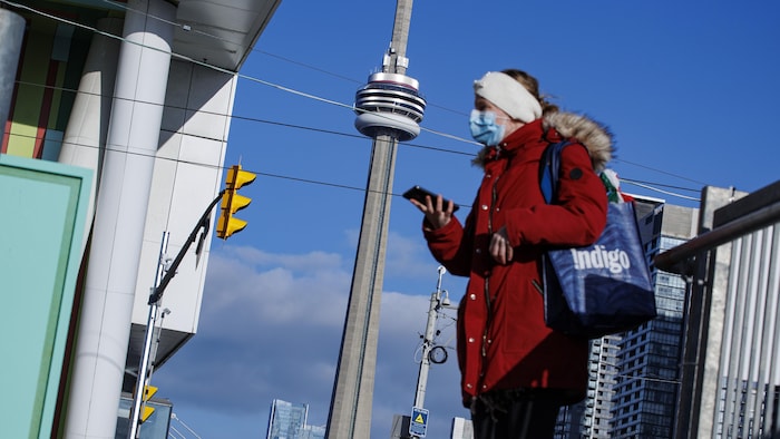 Une femme qui porte un masque sanitaire marche dans une rue à Toronto. La Tour du CN se trouve à l'arrière-plan.