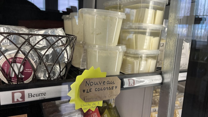 Vue rapprochée d'un réfrigérateur commercial présentant divers produits laitiers. On y voit du beurre emballé, des pots de fromage cottage nommés « Le Colosse » et du fromage dans le vinaigre, accompagnés d'étiquettes de prix et promotionnelles.