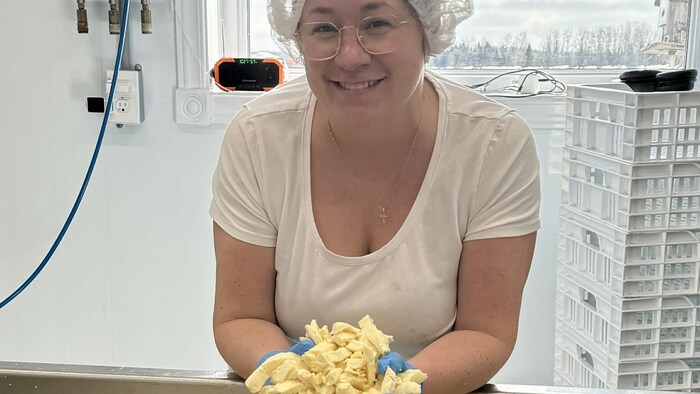 Évelyne Rancourt pose avec du fromage en grains frais sorti d'une cuve. Elle porte des gants et un filet à cheveux.
