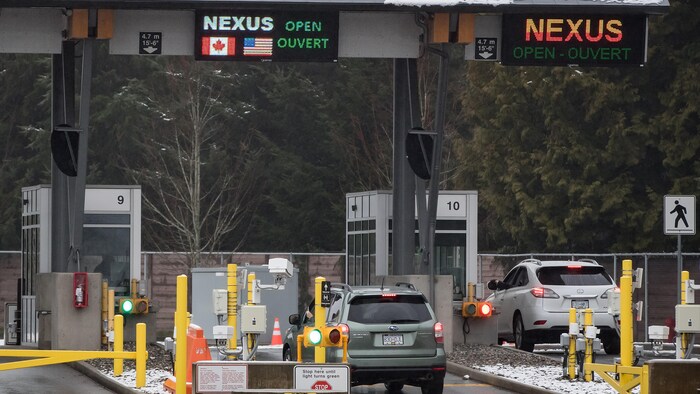Deux voitures traversent la frontière entre le Canada et les États-Unis en hiver. On voit un automobiliste qui passe une carte sur un lecteur optique pour entrer. Au-dessus du poste douanier se trouve la mention « Nexus Open Ouvert ».