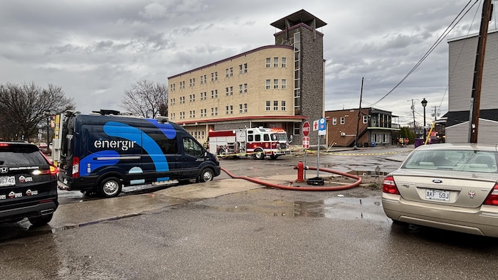 Un coin de rue où une camionnette de compagnie de gaz et un camion de pompier sont stationnés. Un boyau de pompier est branché à une borne d'incendie.