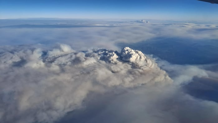 Vue aérienne depuis un hublot d'avion. Une épaisse couche de fumée grise recouvre le paysage jusqu'à l'horizon. L'aile de l'appareil apparaît en haut de l'image, sous un ciel bleu dégagé.