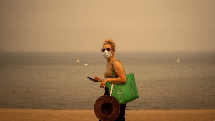 Une femme masquée marche devant une plage extrêmement enfumée.