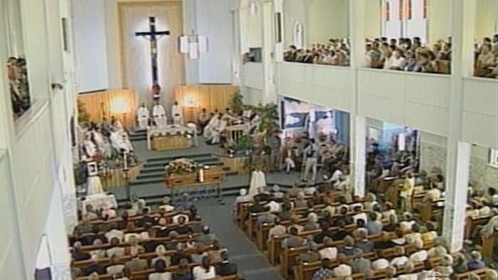 Une église est bondée de personnes. Elles sont assises sur les bancs en bois. Devant elles, un curé célèbre les funérailles d'une personne. 