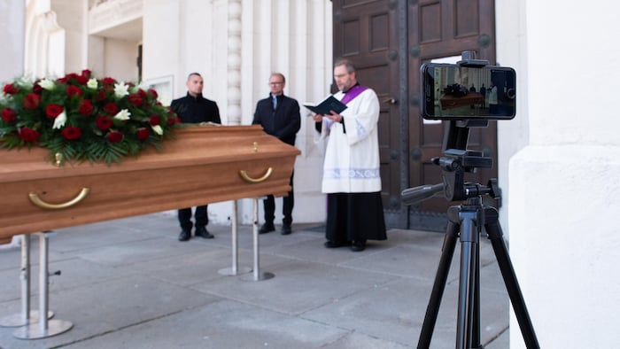 Un prêtre parle devant un cercueil en étant filmé par un téléphone cellulaire sur trépied.