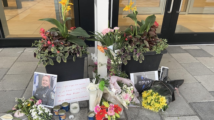 Un mémorial improvisé comprenant une photo du policier et quelques bouquets de fleurs, par terre devant un commerce.