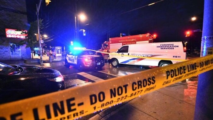 Une scène de crime, la nuit, entourée de ruban jaune de police. Plusieurs véhicules d'urgence sont sur place.