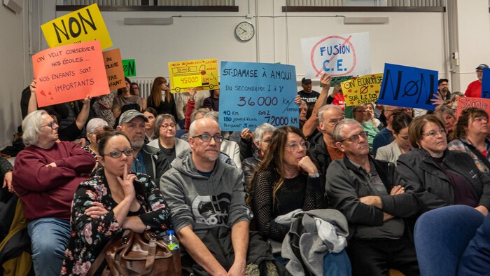 Des gens assis dans une salle tiennent des pancartes aux slogans revendicateurs.