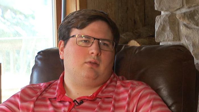 Un jeune homme vêtu d'un polo rayé couleur saumon est assis dans un fauteuil brun en cuir.