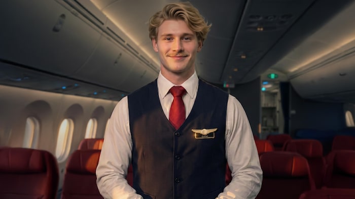 Natan-Maël Gray en uniforme d'agent de bord, pose au centre de la cabine d'un avion commercial dont les rangées de sièges sont inoccupées.