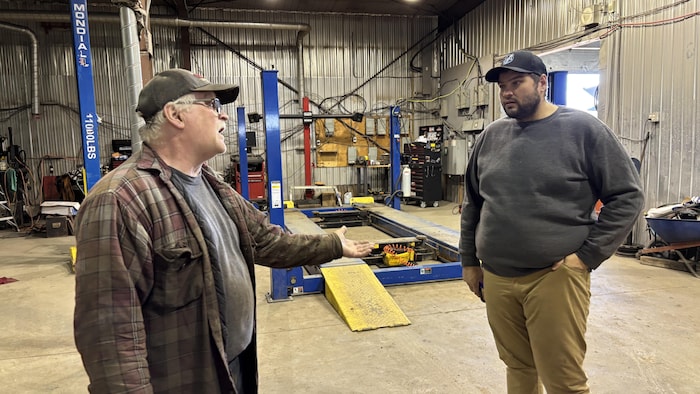 Deux hommes, un plus âgé portant une casquette et une chemise à carreaux gesticulant de la main, et un plus jeune écoutant attentivement, discutent dans l'atelier mécanique.
